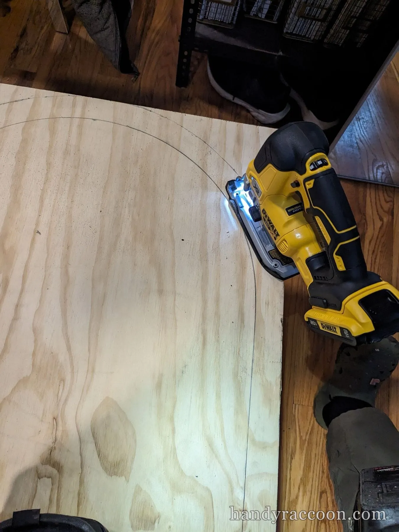 A yellow cordless jigsaw following a pencil curve across a plywood sheet on the floor.