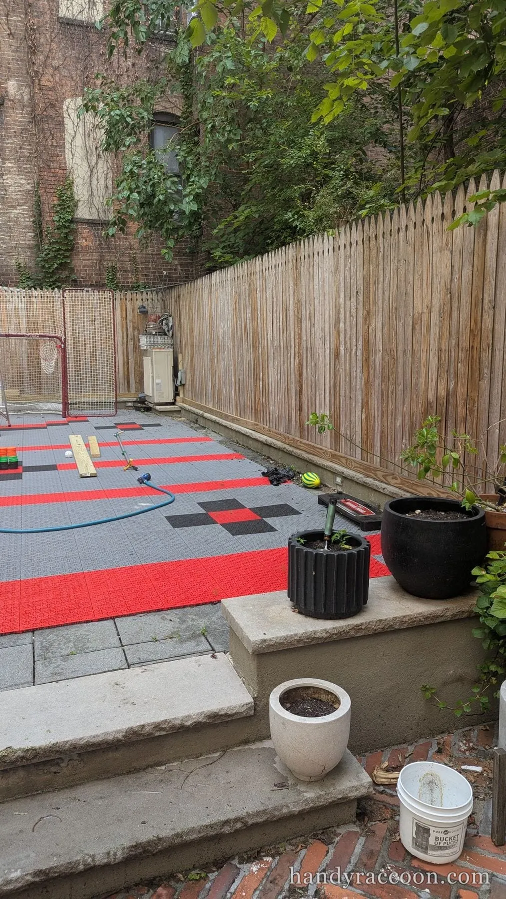 A backyard with red and gray sport tiles, a hockey net, potted plants and a wood fence.