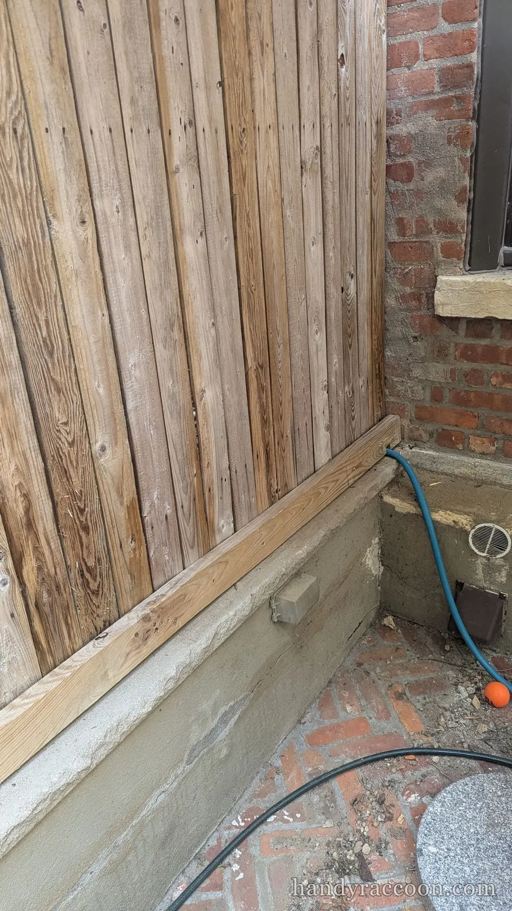 A fresh light board installed along the bottom of a gray wood fence beside a brick wall.