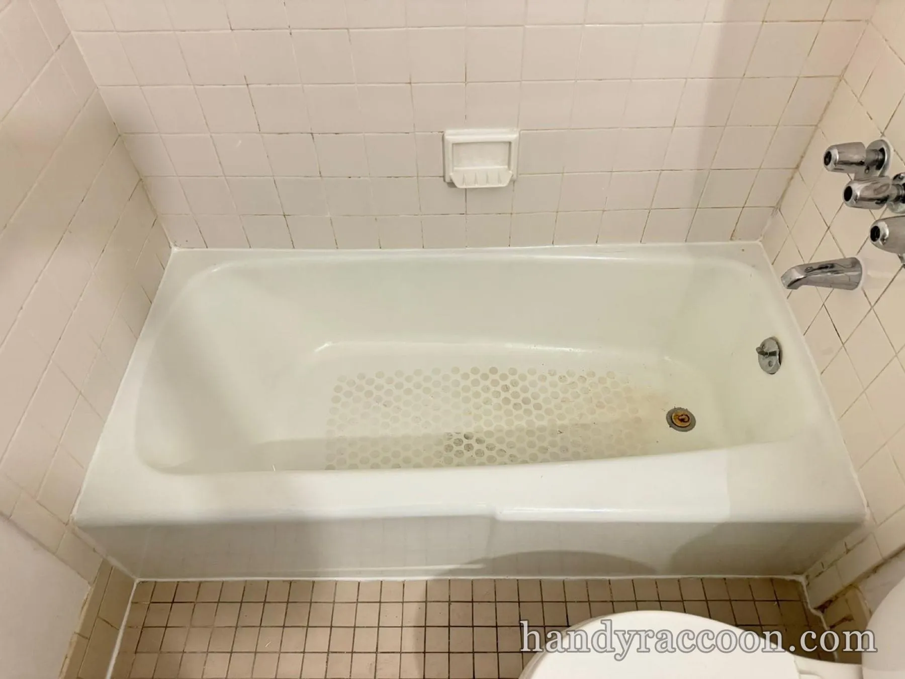 Older white bathtub with stained grout, square wall tile and a beige tile floor.