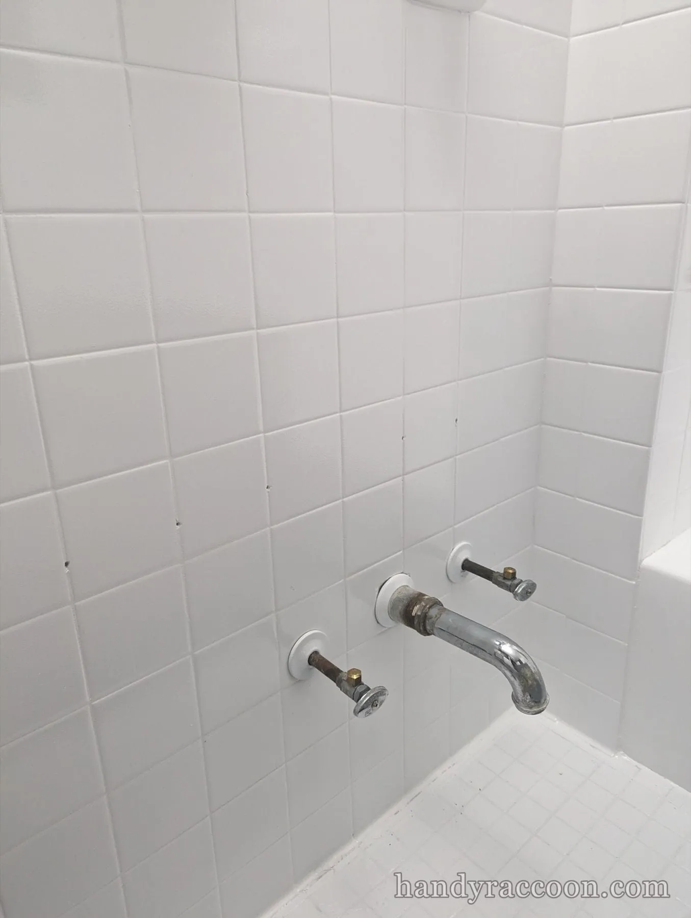 White square tile bathtub wall with a chrome spout and two exposed valve stems.