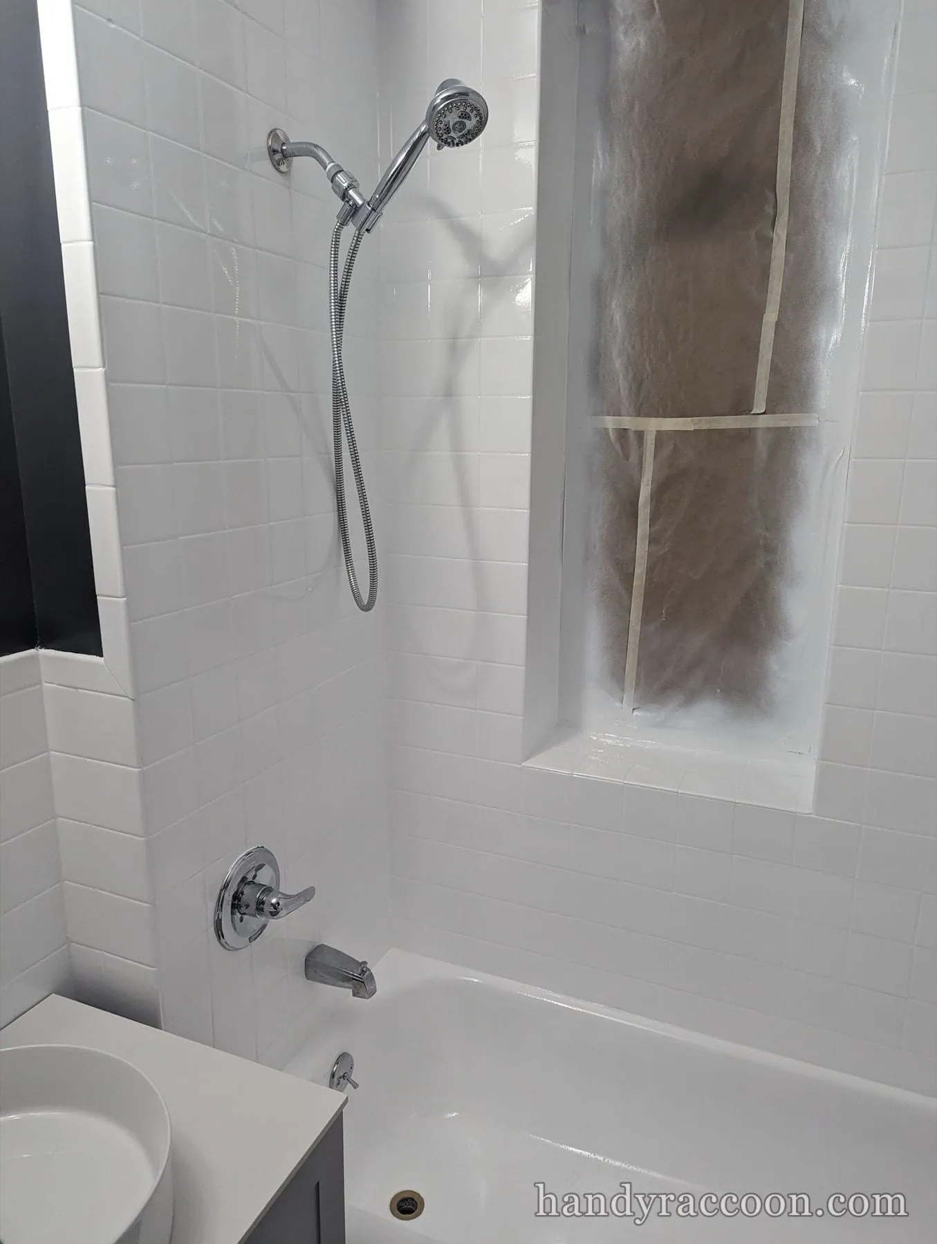 White tile tub surround with a handheld shower and a niche covered in masking paper.
