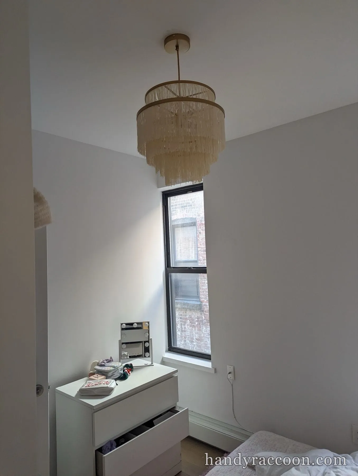 Two-tier cream fringe chandelier mounted on a white bedroom ceiling near a black-framed window.