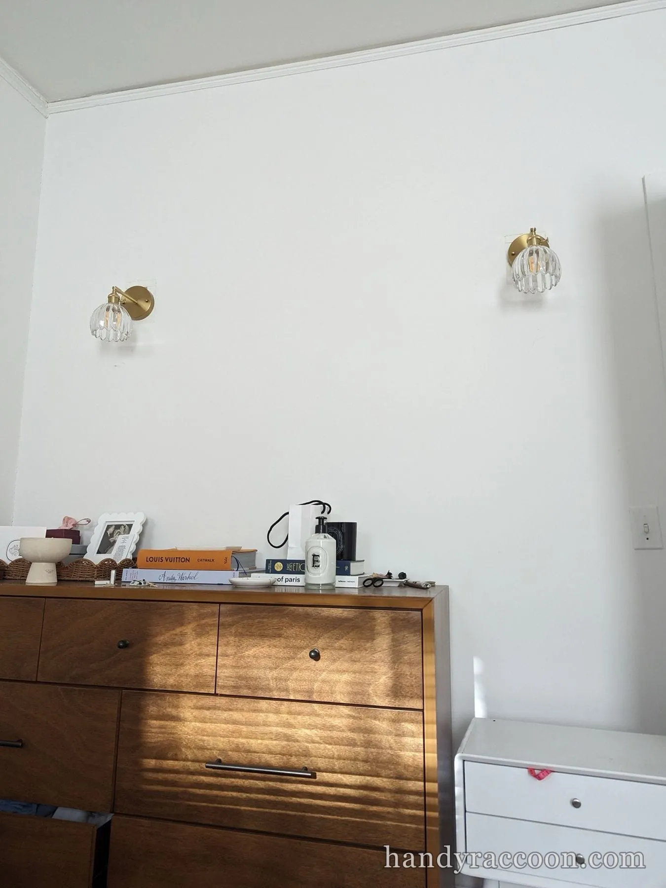 Two clear glass and brass wall sconces installed on a white wall above a wooden dresser.