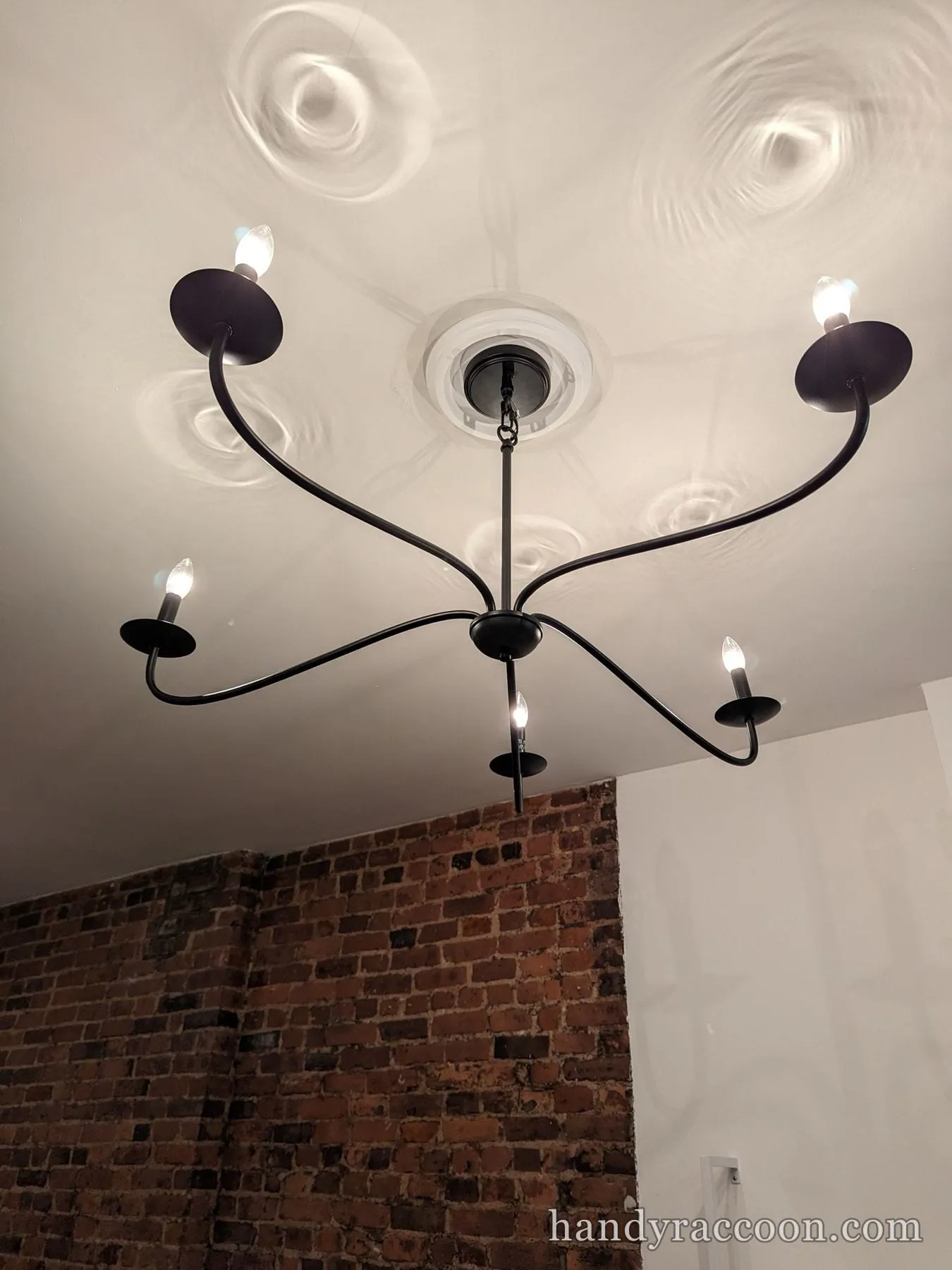 Black curved five-arm candle chandelier lit against a white ceiling and brick wall.
