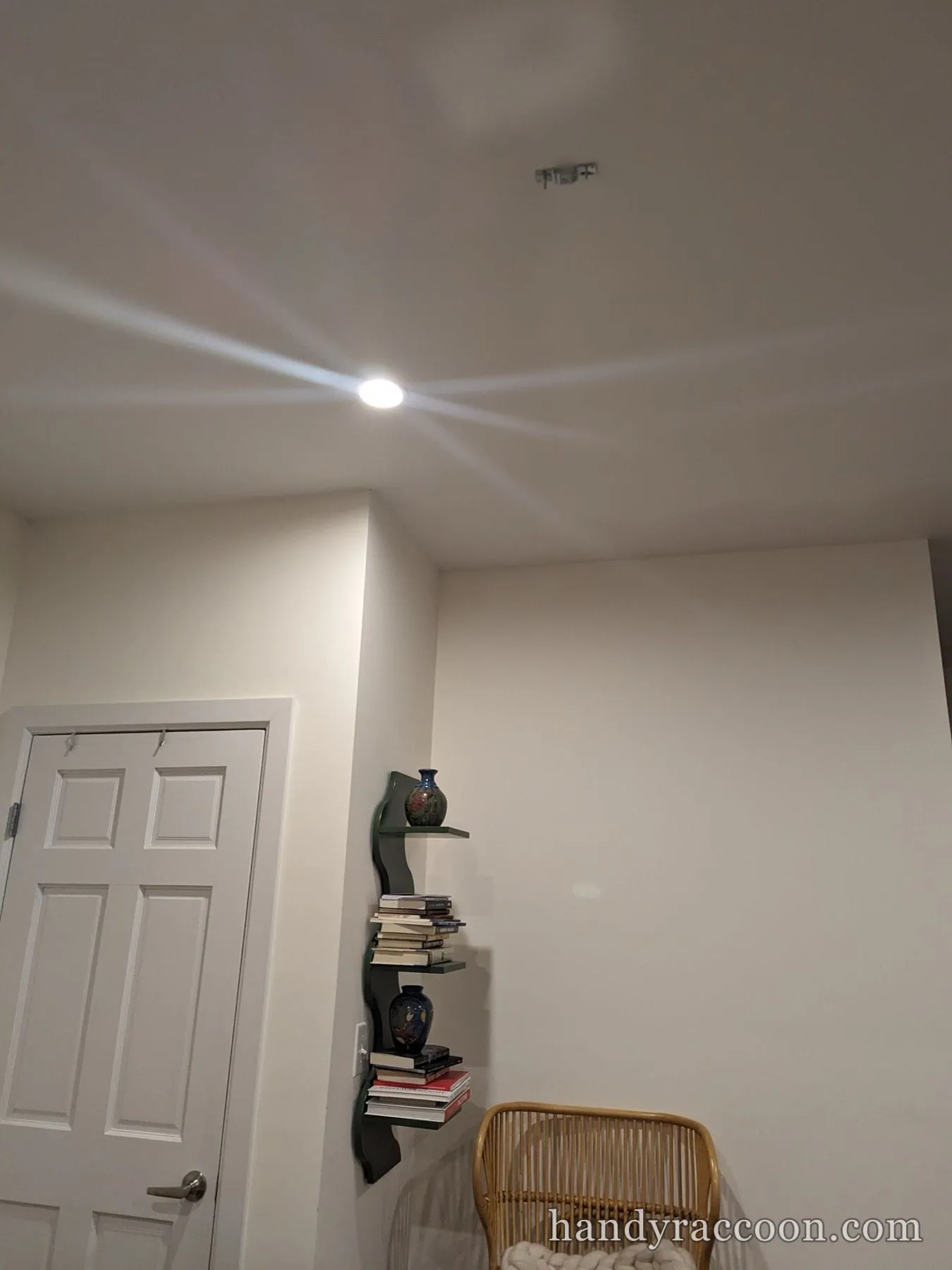 Lit recessed ceiling downlight throwing a star flare in a room with a door and corner book shelf.