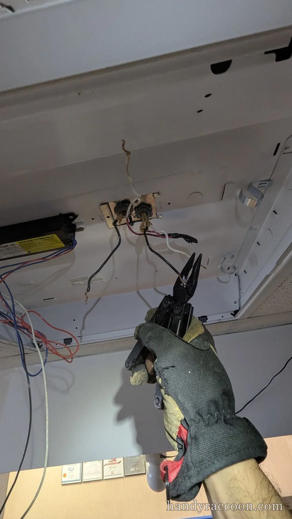 Gloved hand with pliers working on wiring inside an opened fluorescent ceiling fixture.