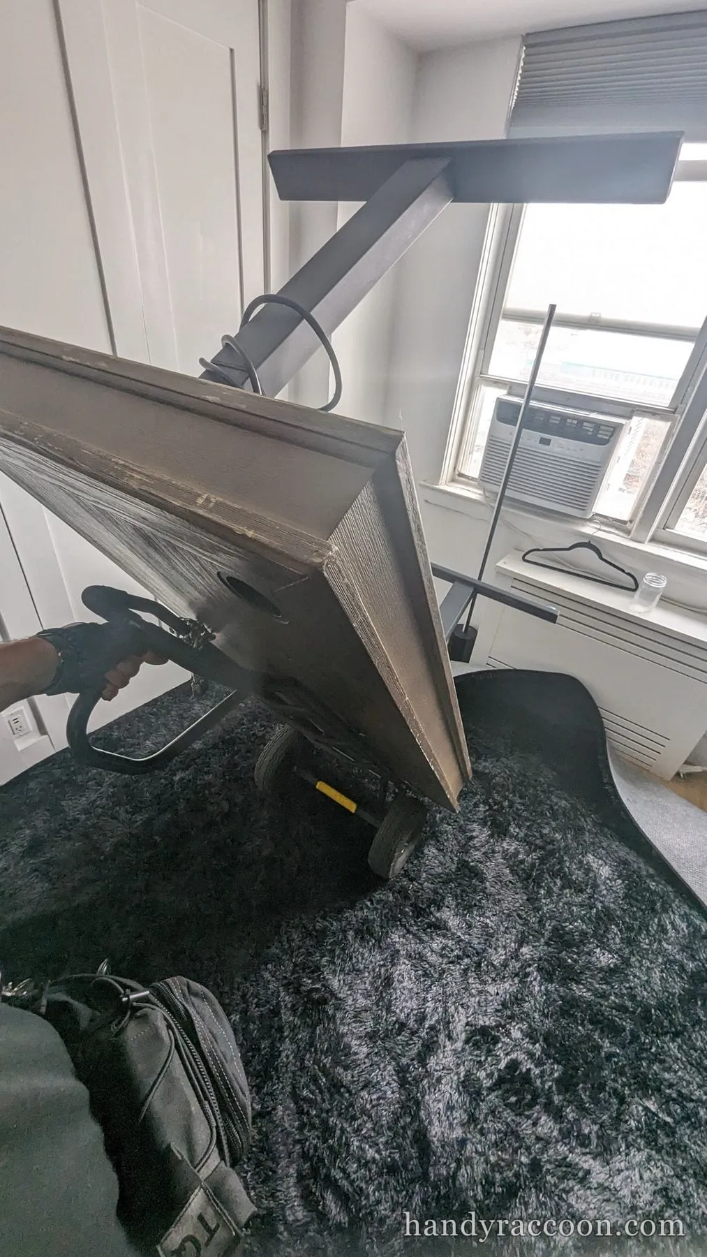 A gloved hand shrink-wrapping a grey cabinet tilted back on a hand truck in a carpeted room.