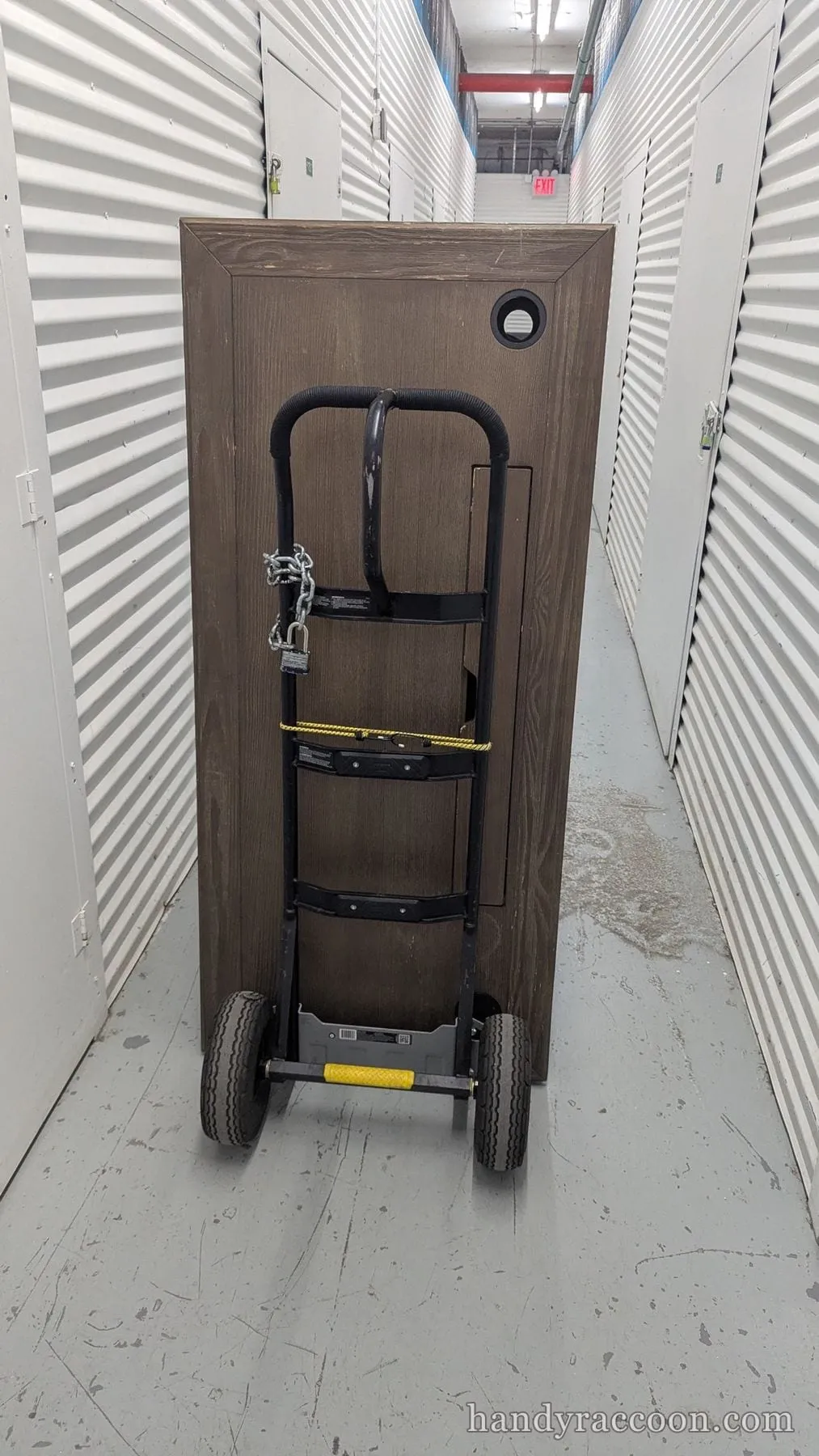 A tall wood-grain cabinet strapped upright to a hand truck in a storage unit hallway.
