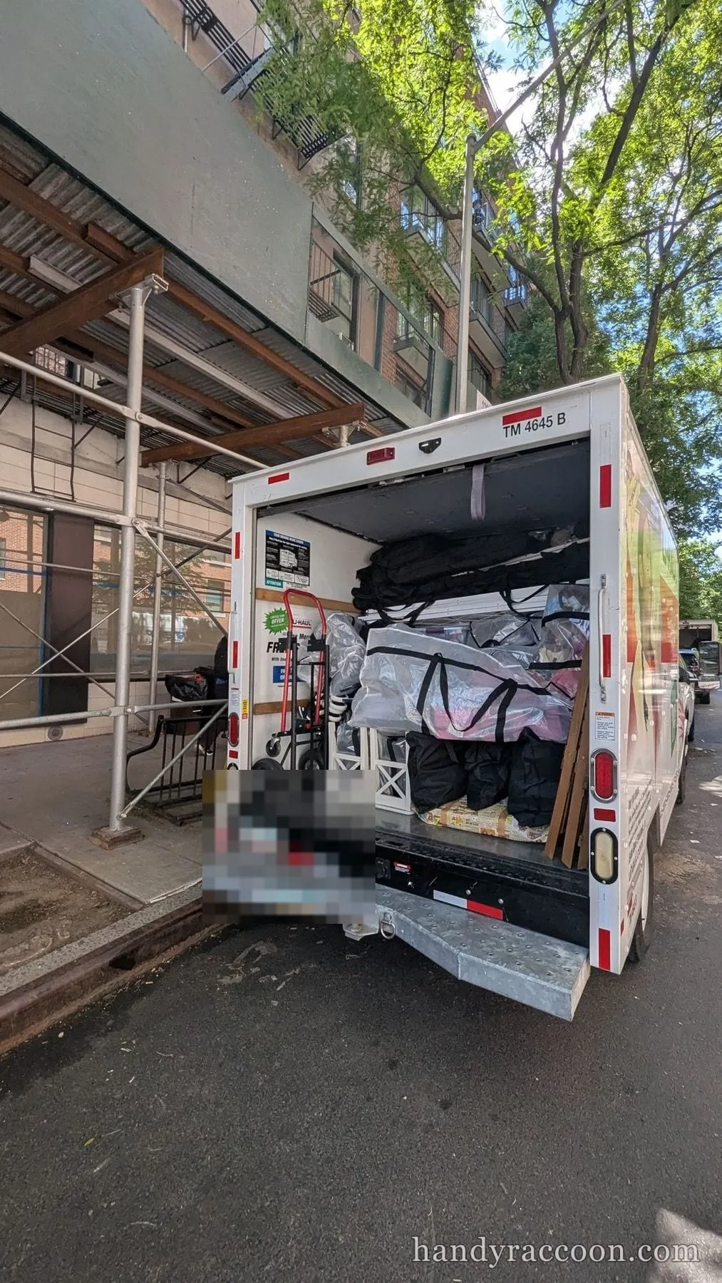 Rear of an open box truck on a city street loaded with plastic-wrapped furniture, bags, and a hand truck.