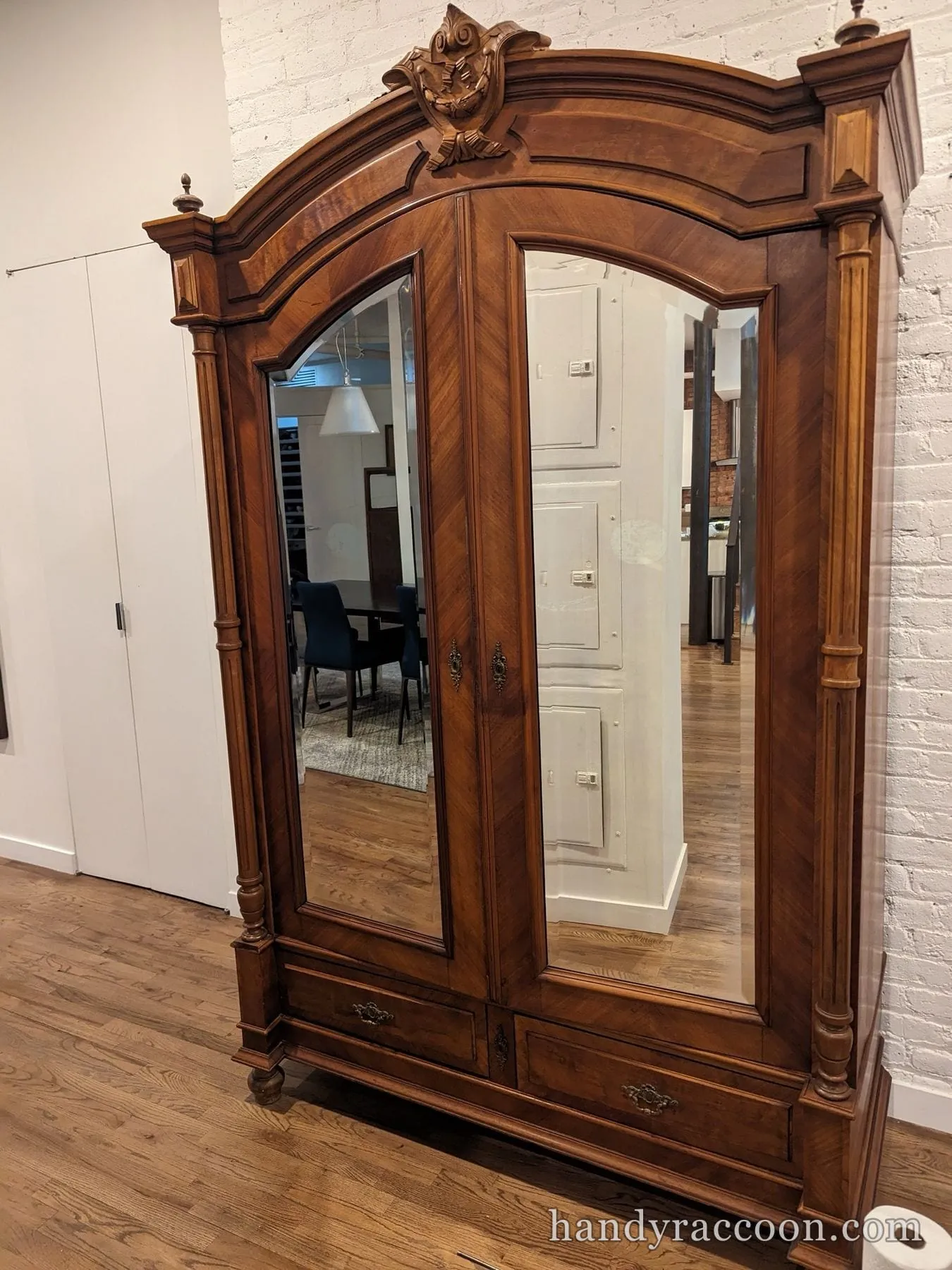 Tall carved antique wood wardrobe with two beveled mirror doors by a brick wall.