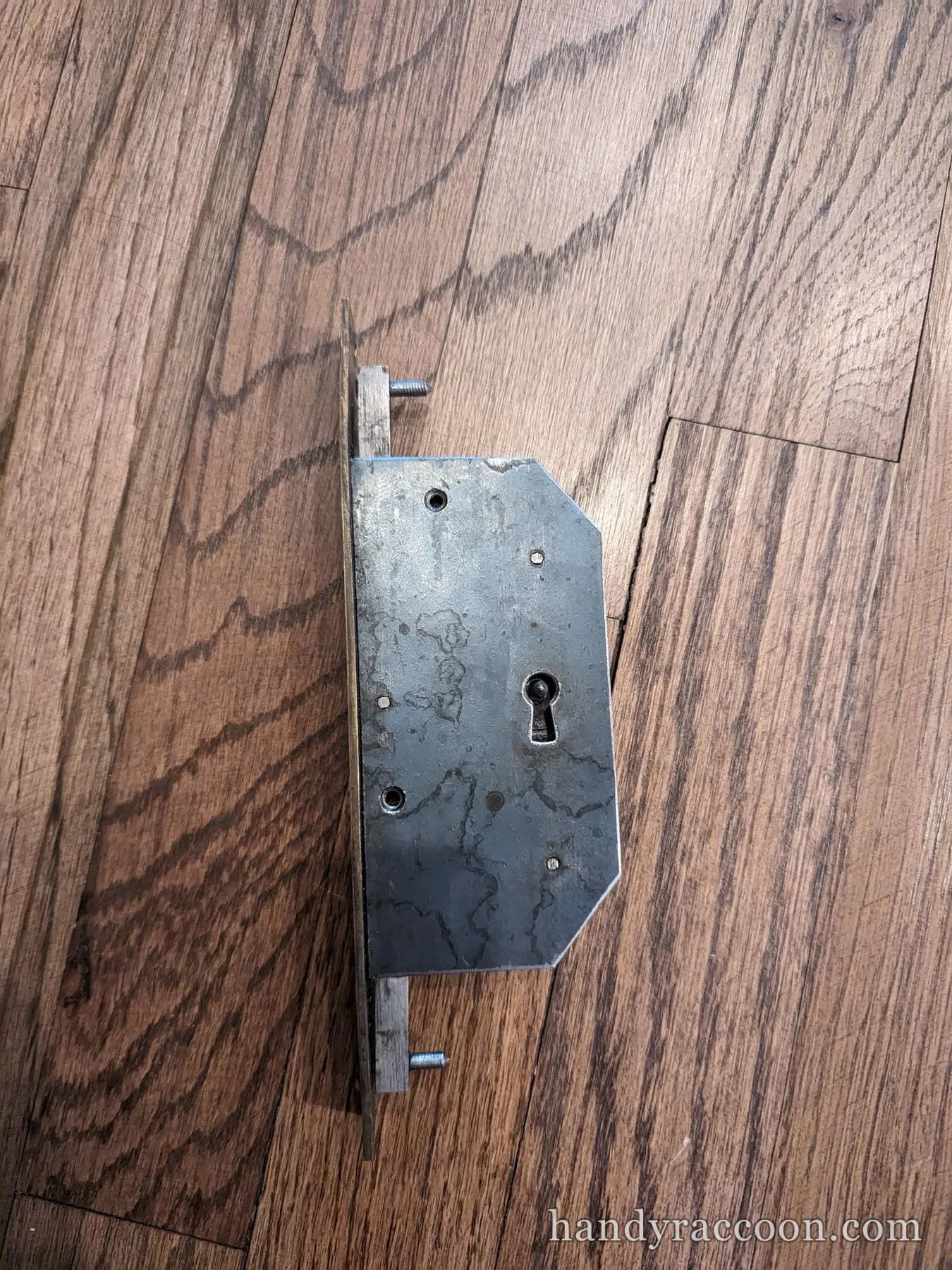 An old skeleton-key mortise lock body resting face-up on a hardwood floor.