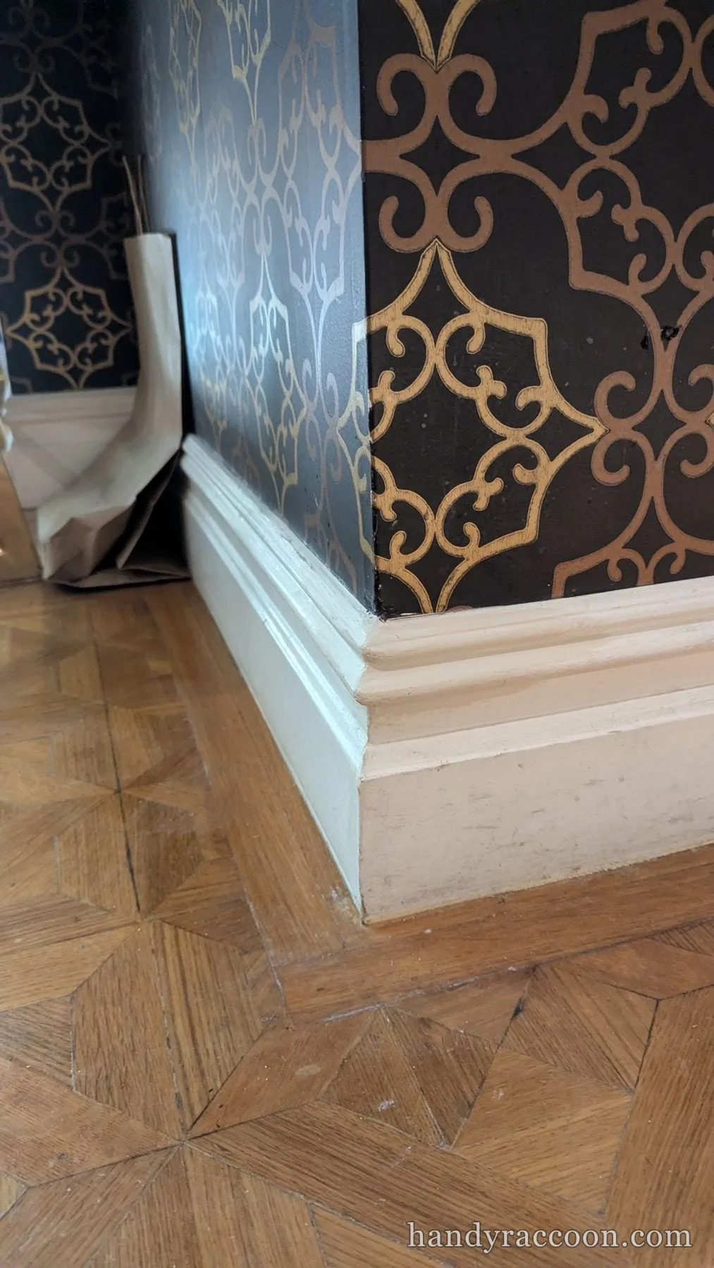 Repaired baseboard corner, filled and primed, next to black and gold patterned wallpaper on parquet floor.