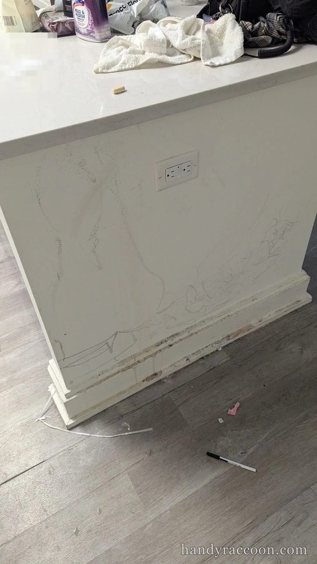 White kitchen island end panel with pencil scribbles and water-stained baseboard, cluttered counter above.
