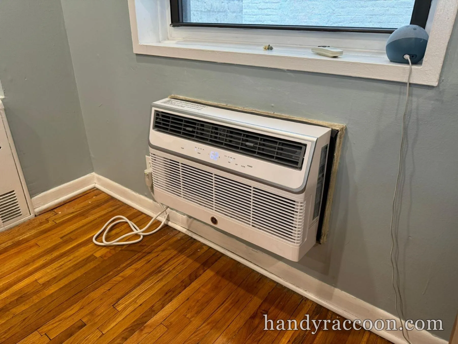 White GE through-wall air conditioner mounted in a wall sleeve below a window over a wood floor.