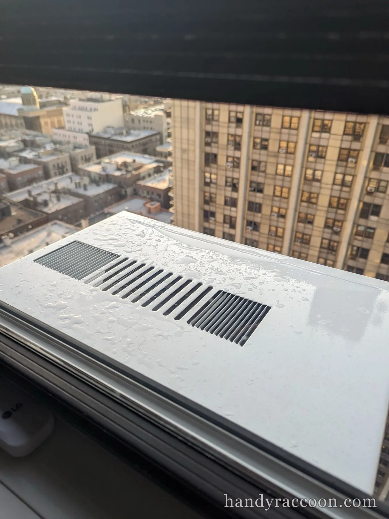 Top of a white window air conditioner beaded with water, with high-rise buildings beyond the window.