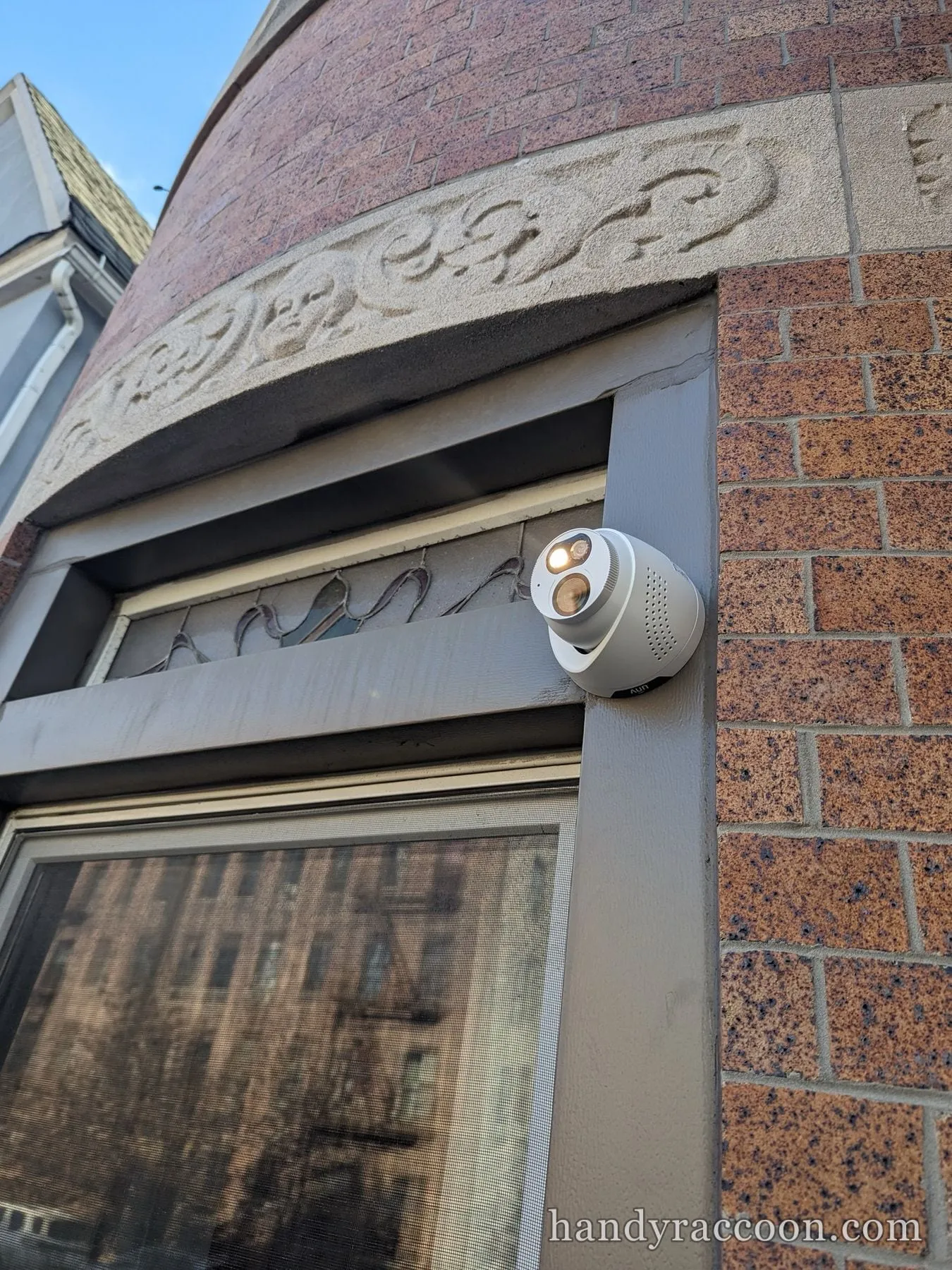 White dome security camera with an illuminated spotlight installed on a brick building corner below decorative stone trim.