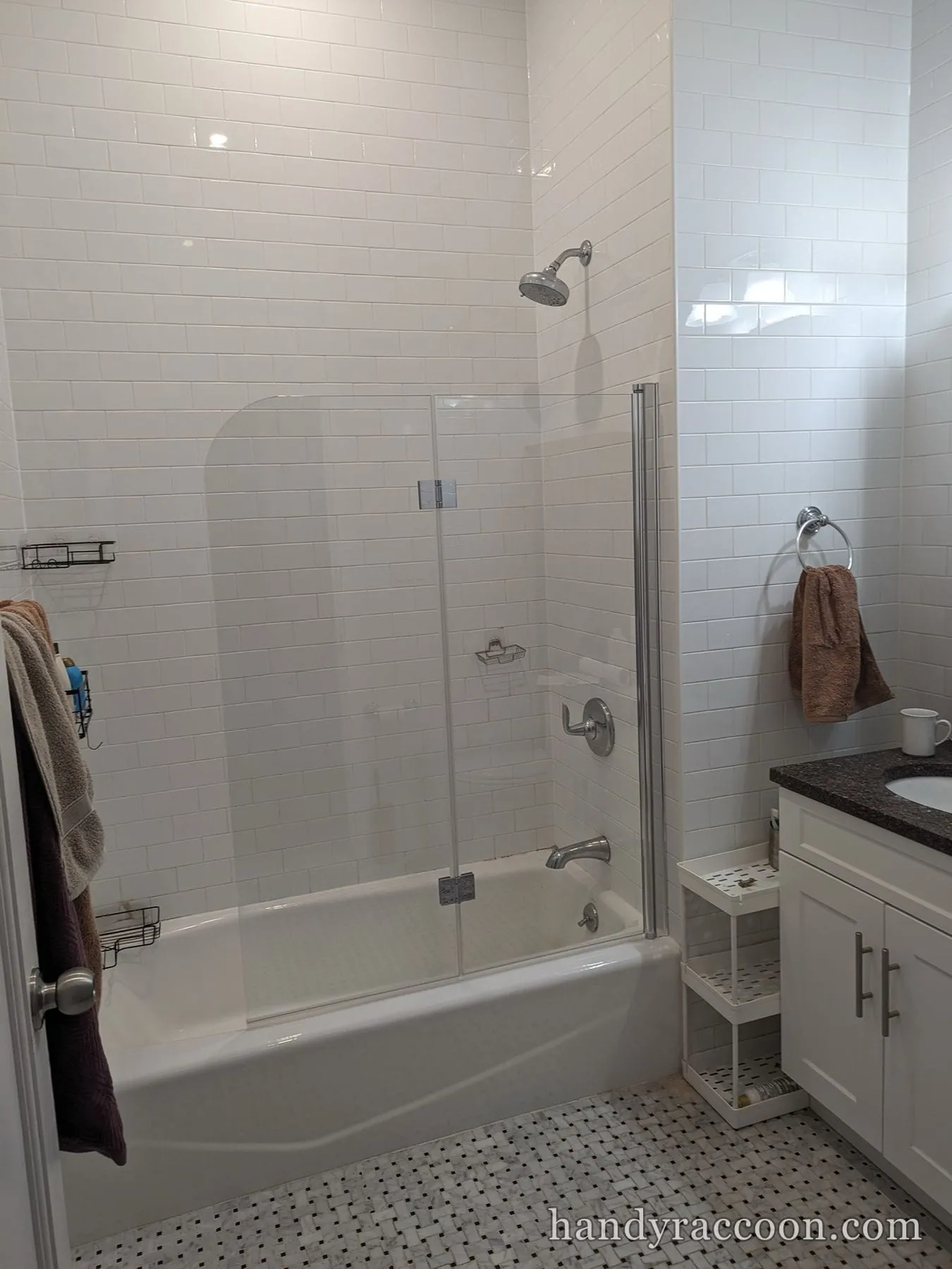 Folding clear glass shower door on a white bathtub with white subway tile, stone-top vanity and towel ring.