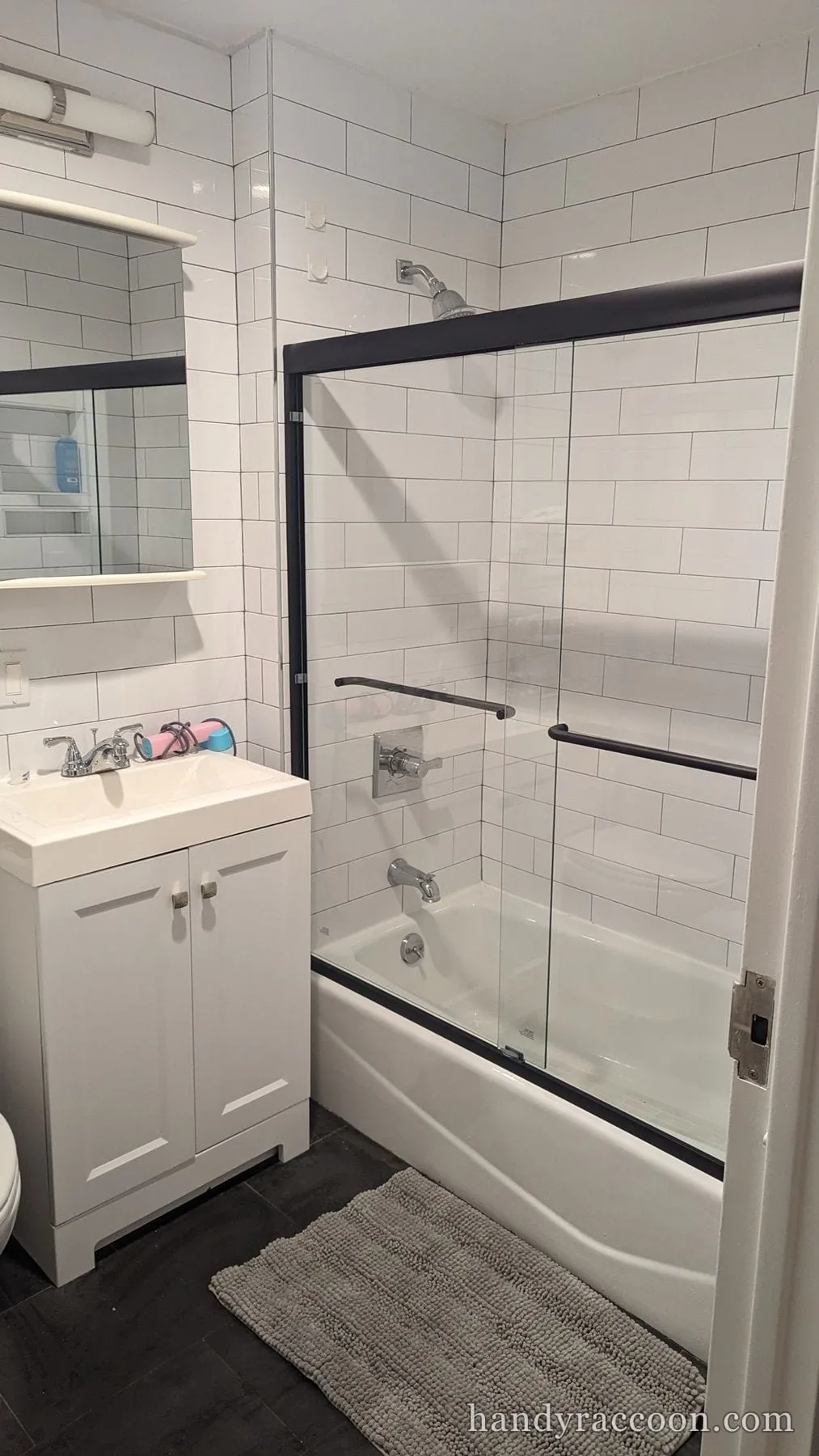 Black-framed sliding glass shower doors on a white bathtub with white subway tile, dark floor, and white vanity.