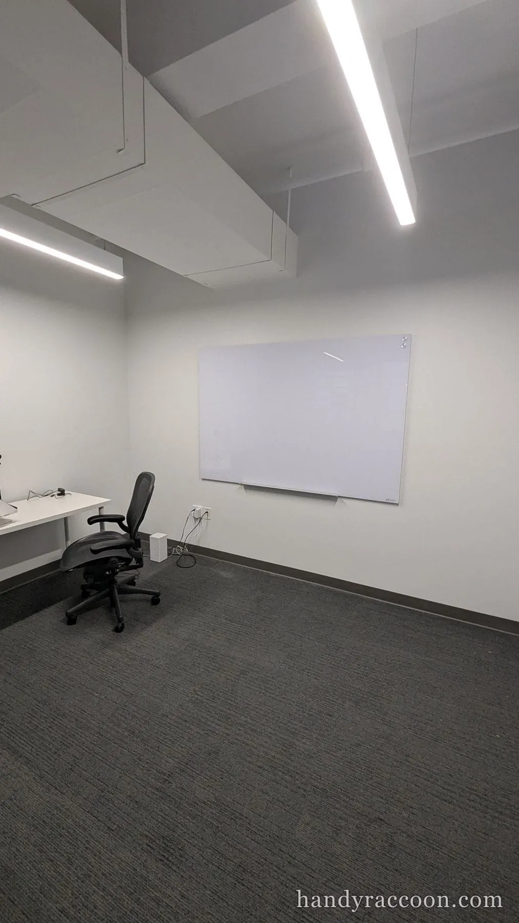 A glass dry-erase board mounted on a gray wall in an empty office with a white desk and black chair.