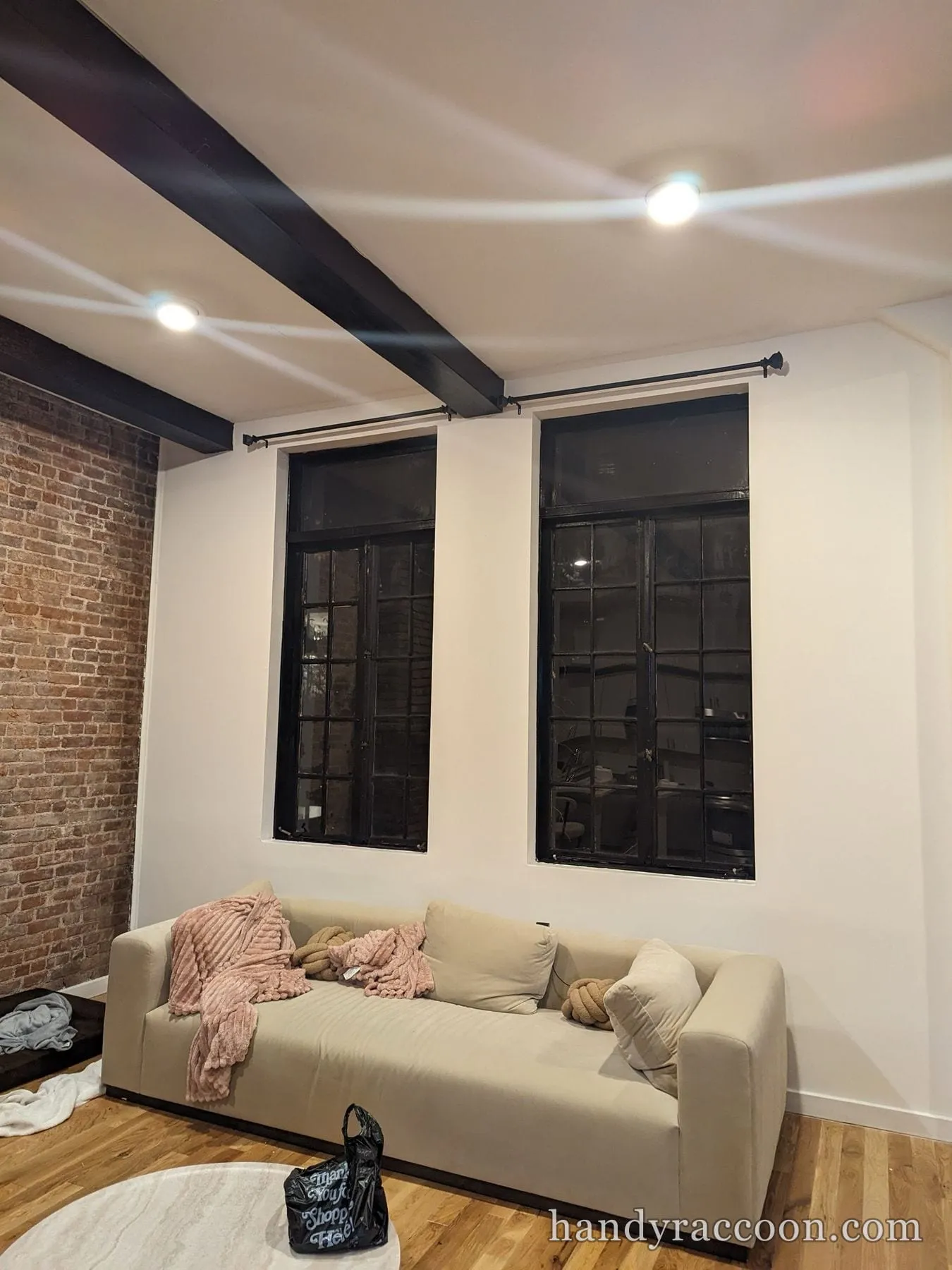 Living room with exposed brick, tan sofa, and two tall black windows with black curtain rods mounted above them.