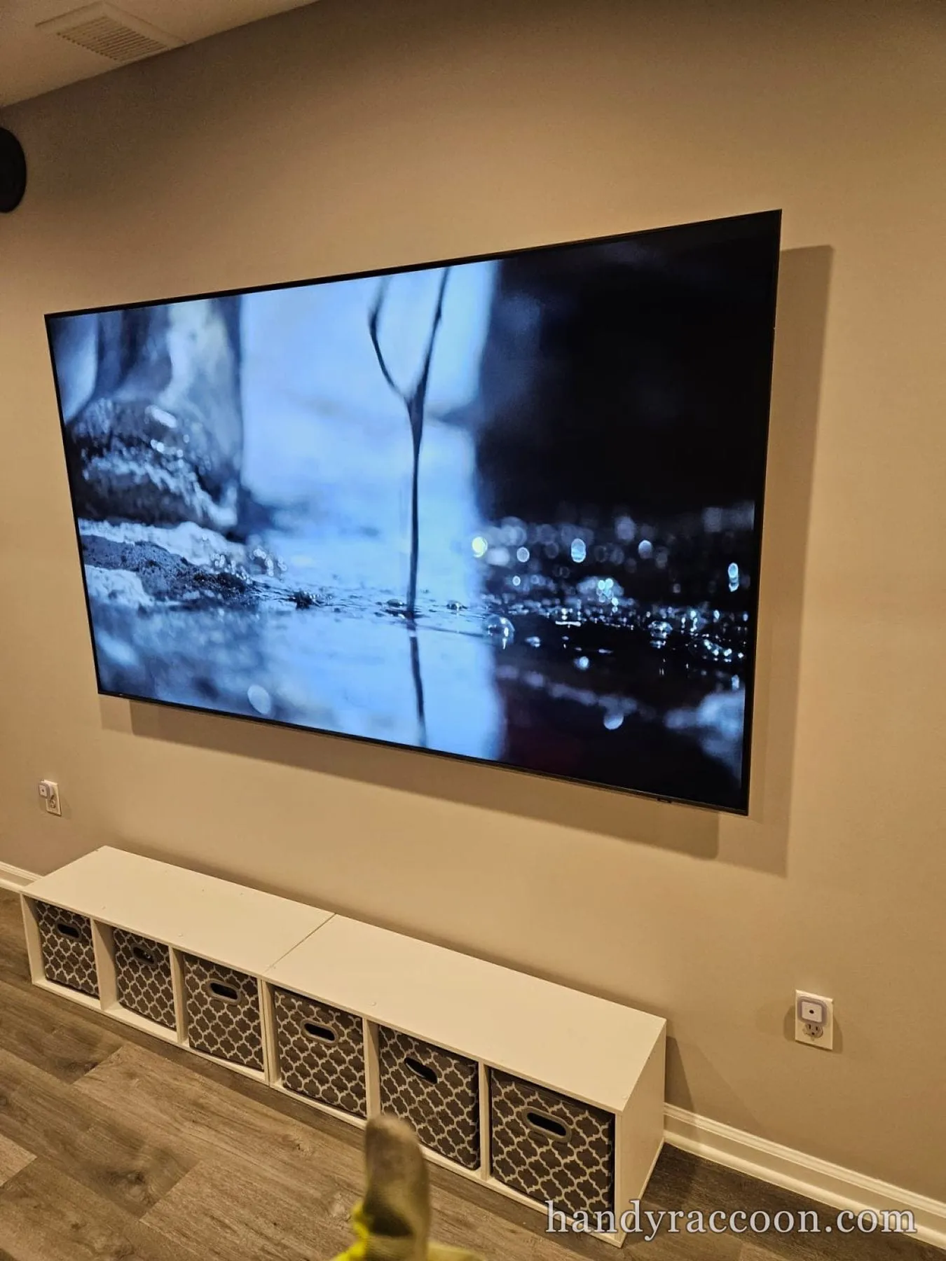 Large flat-screen TV mounted on a tan wall above a white six-cube storage bench in a basement room.