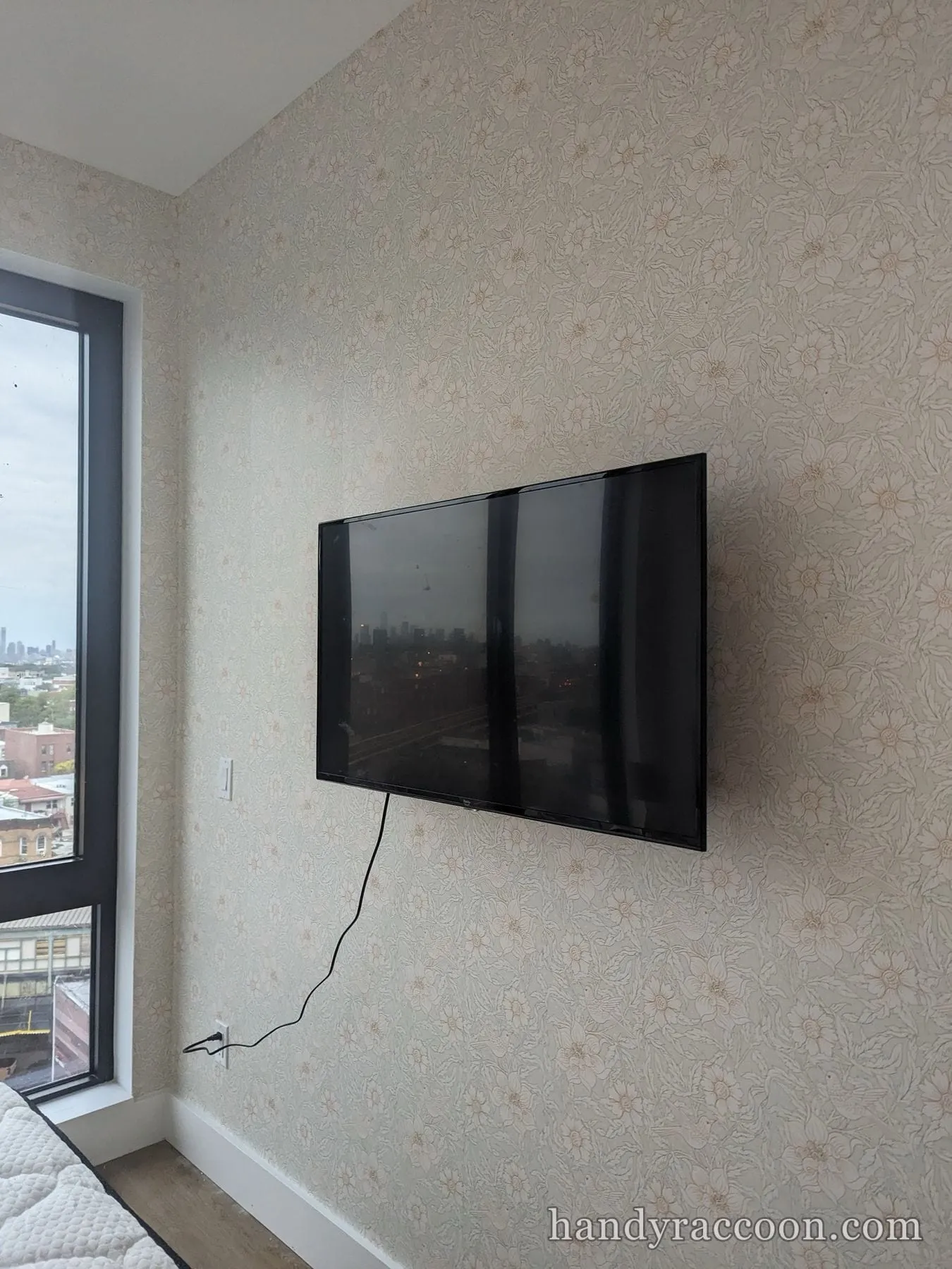 Flat-screen TV mounted on floral-patterned wallpaper next to a window with a city view.