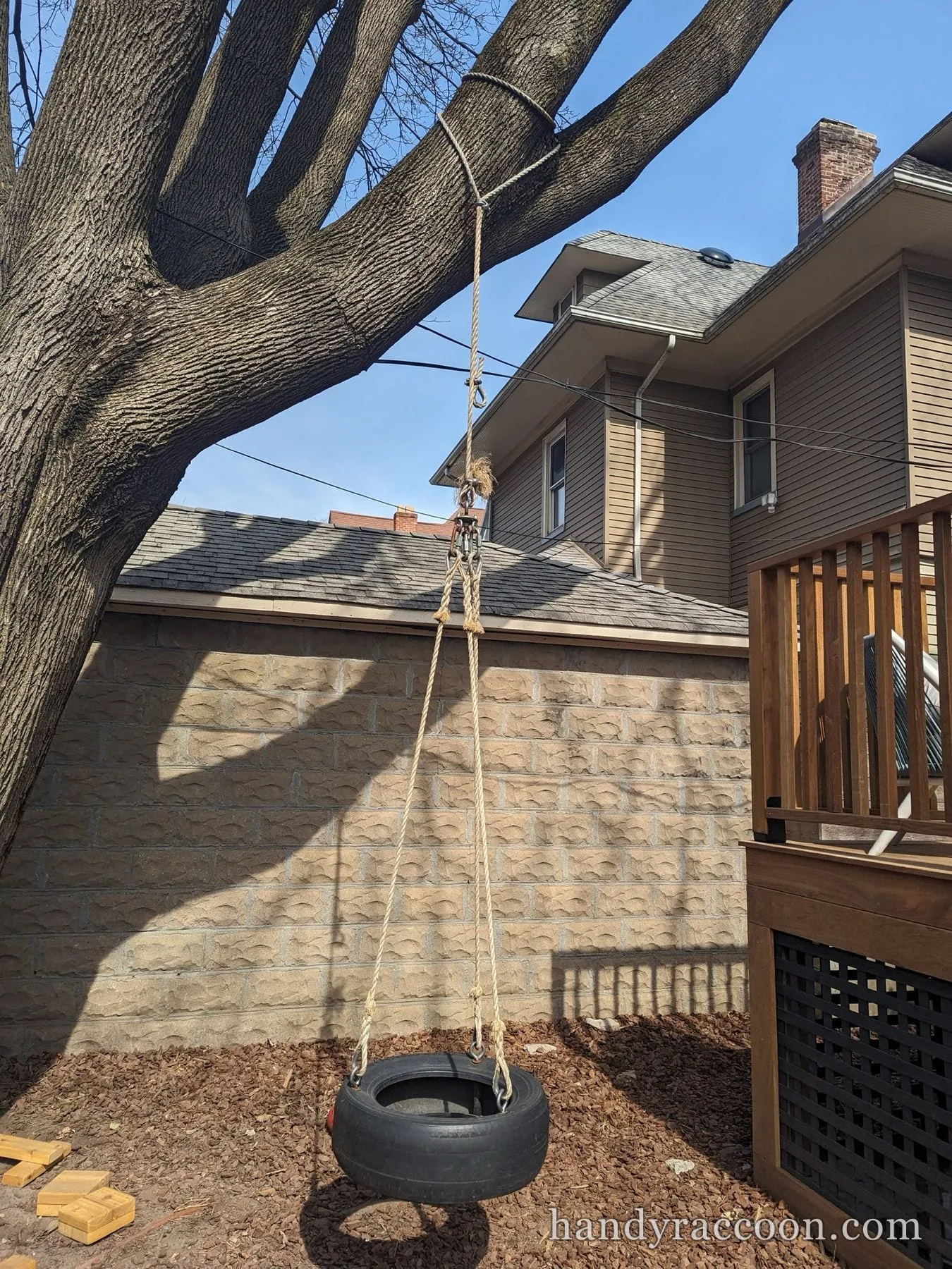 A tire swing hung from a tree branch over a mulch bed beside a wooden deck.