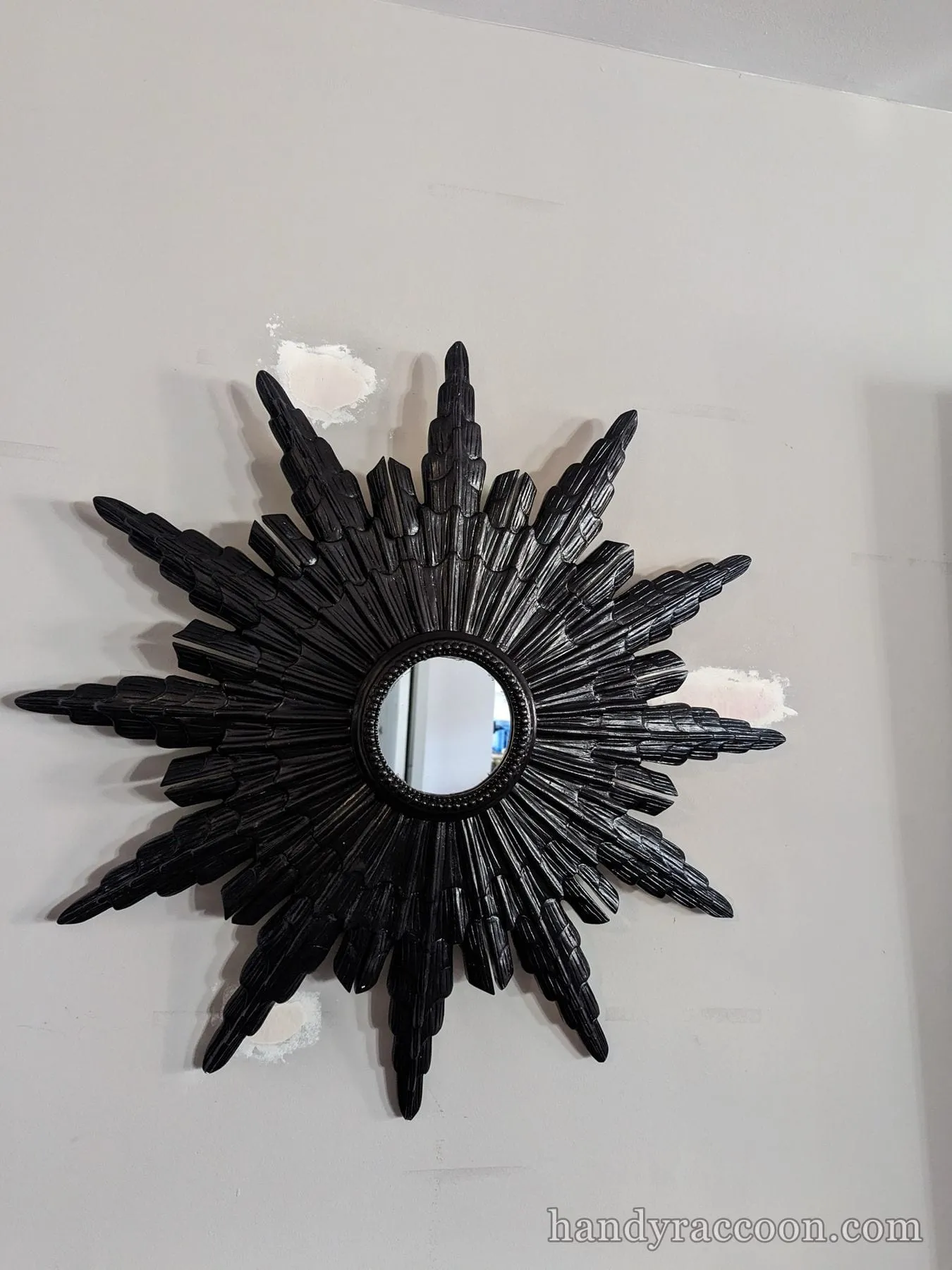 Glossy black sunburst wall mirror mounted on a light wall with visible spackle patches.