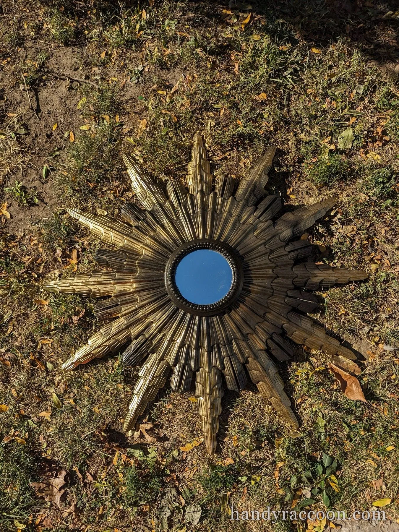 Gold sunburst wall mirror with feathered rays on autumn grass, blue sky reflected in the mirror.