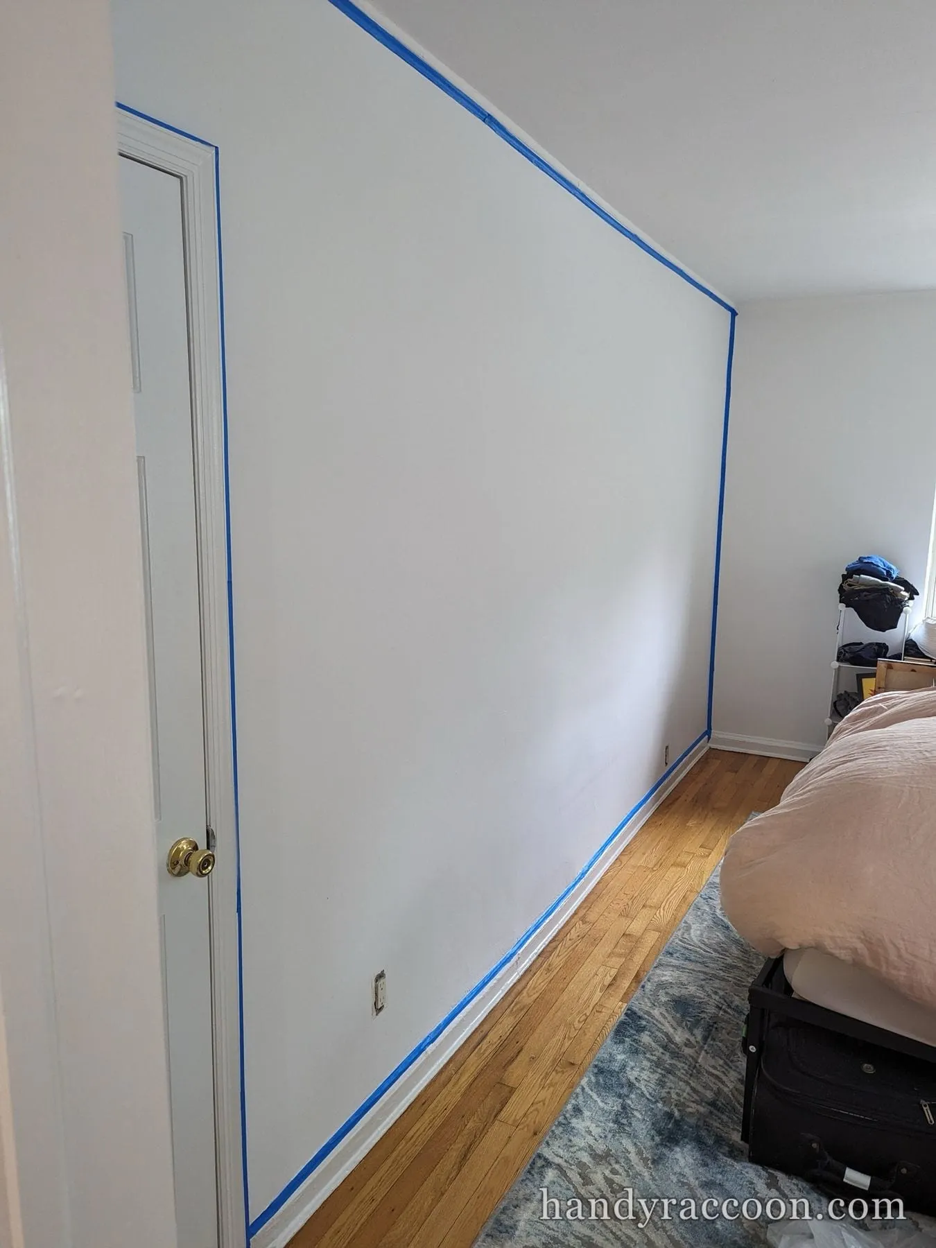 Bedroom wall masked with blue tape along the ceiling, door, and baseboard before painting.