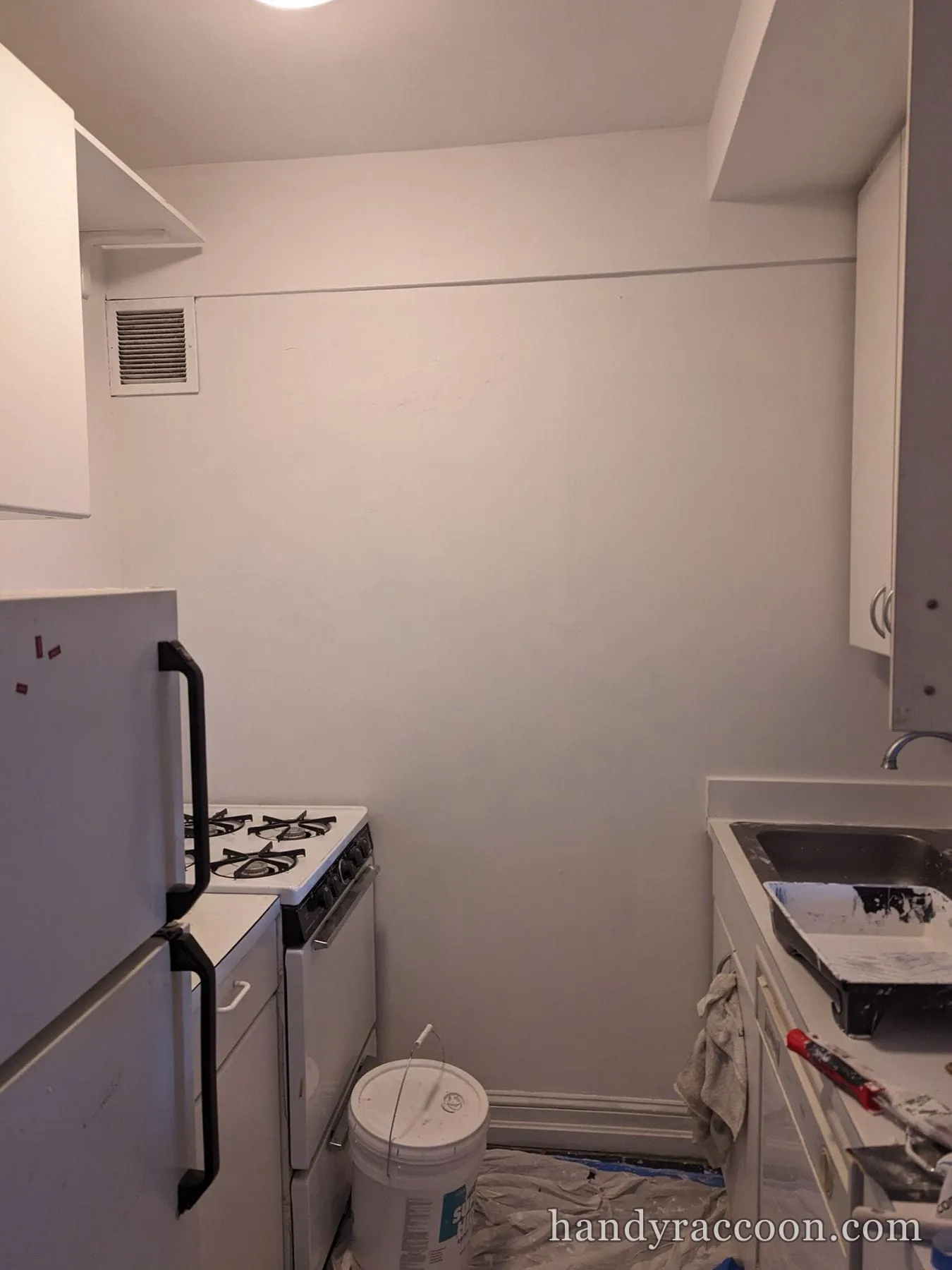 Compact kitchen mid painting with gray walls, drop cloth, bucket and paint tray.