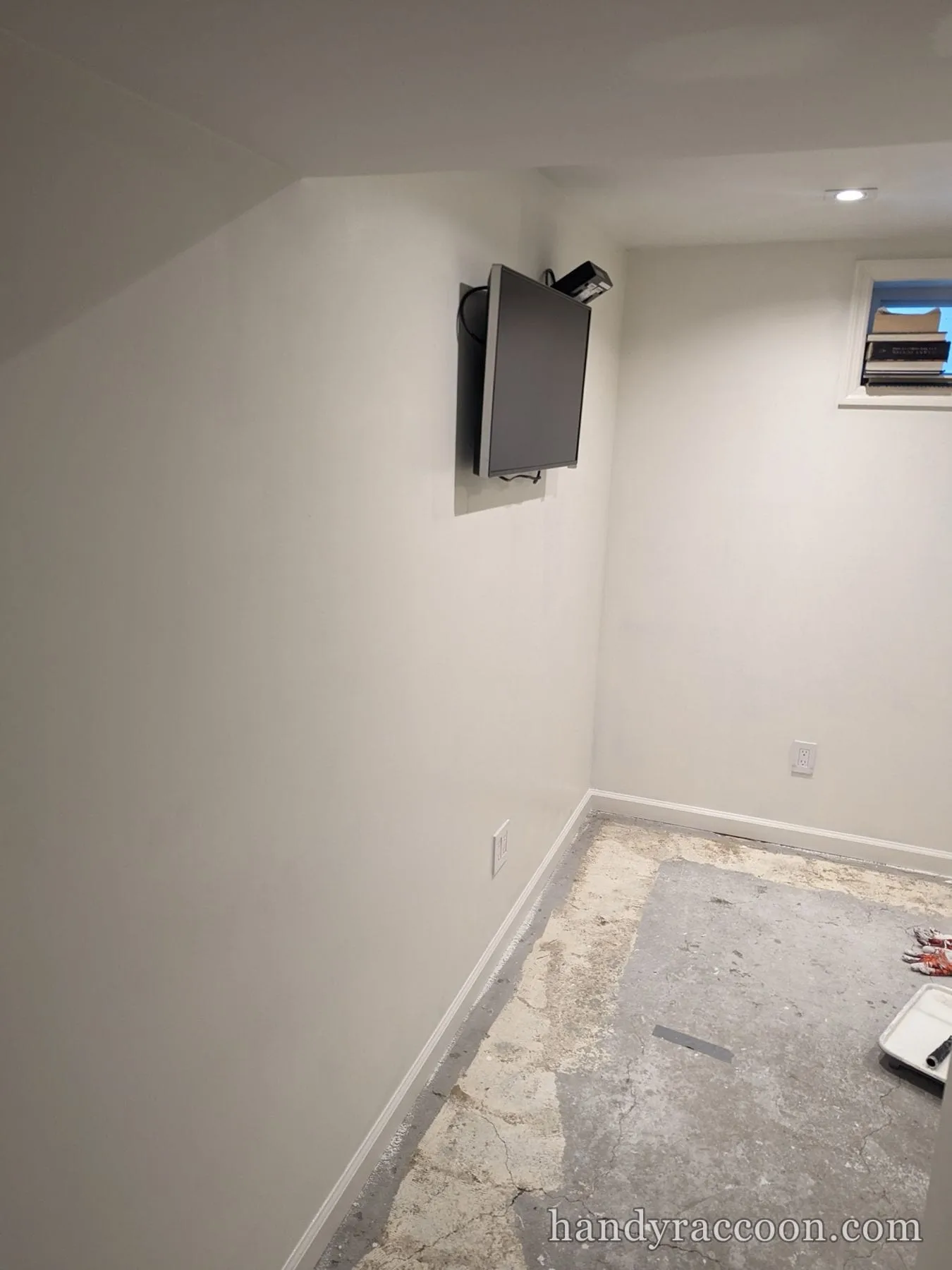 Basement room with freshly painted off-white walls, white baseboards, a wall-mounted TV, and bare concrete floor.