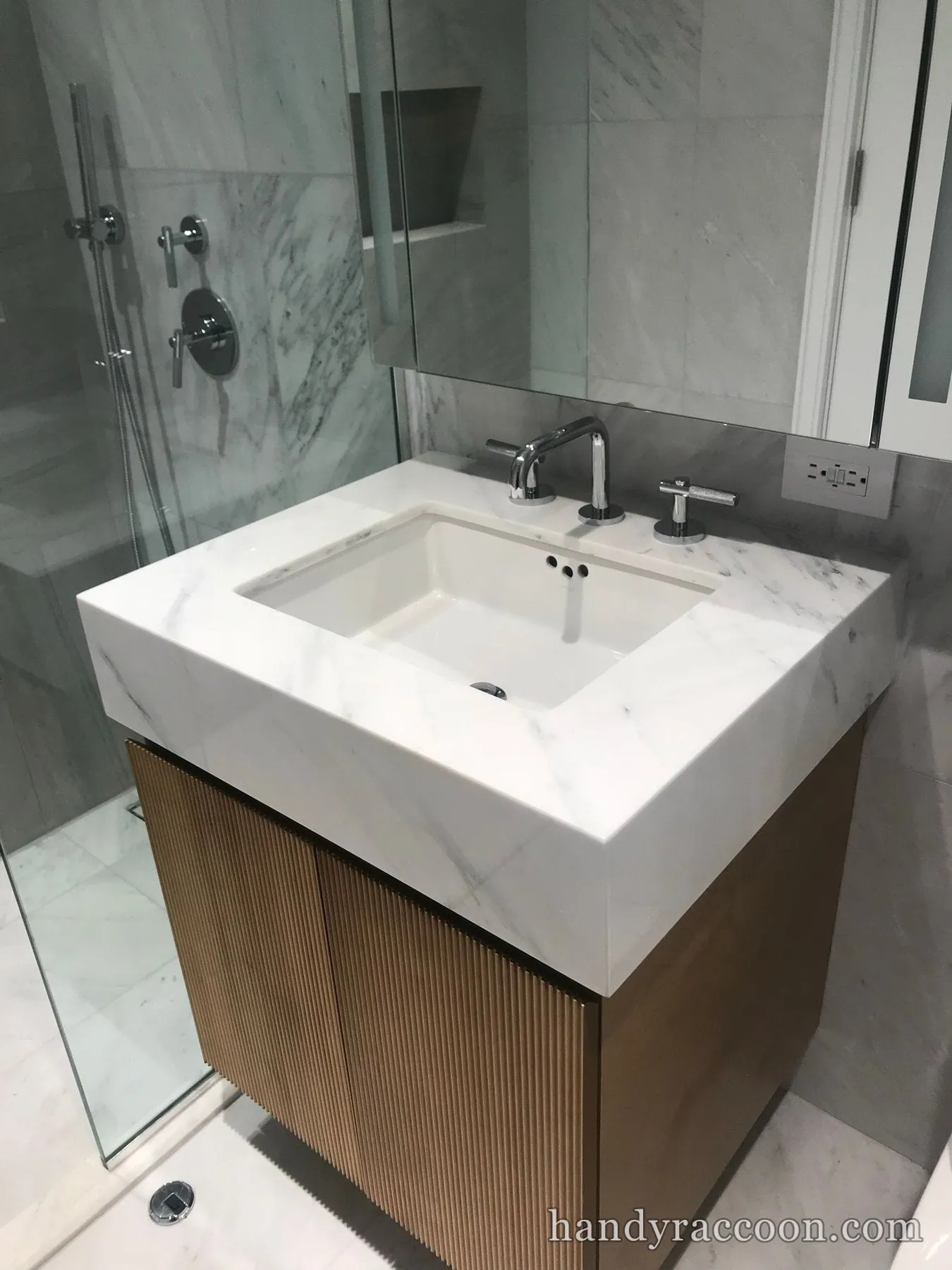 Stone vanity top with undermount sink and chrome faucet, wood fluted cabinet below, glass shower at left.