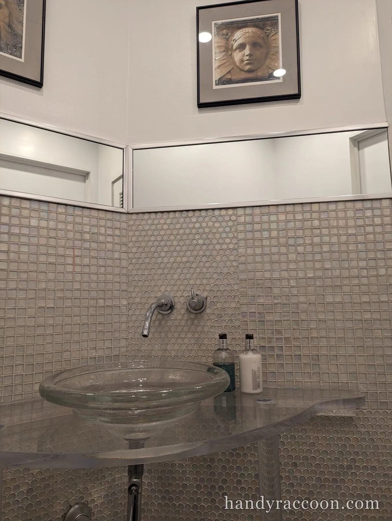 Round glass vessel sink on a glass counter with a chrome wall-mounted faucet and mosaic tile wall.