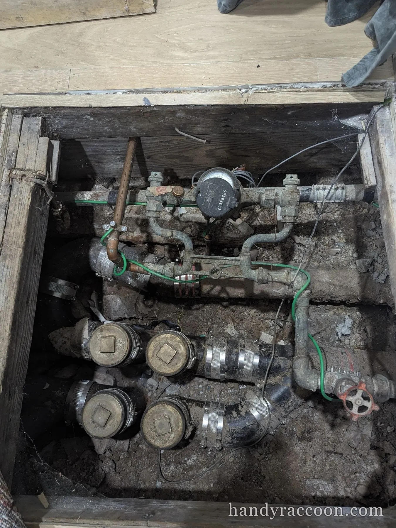 Opened floor hatch revealing a building water meter, valves, and cleanout plugs in a crawlspace.