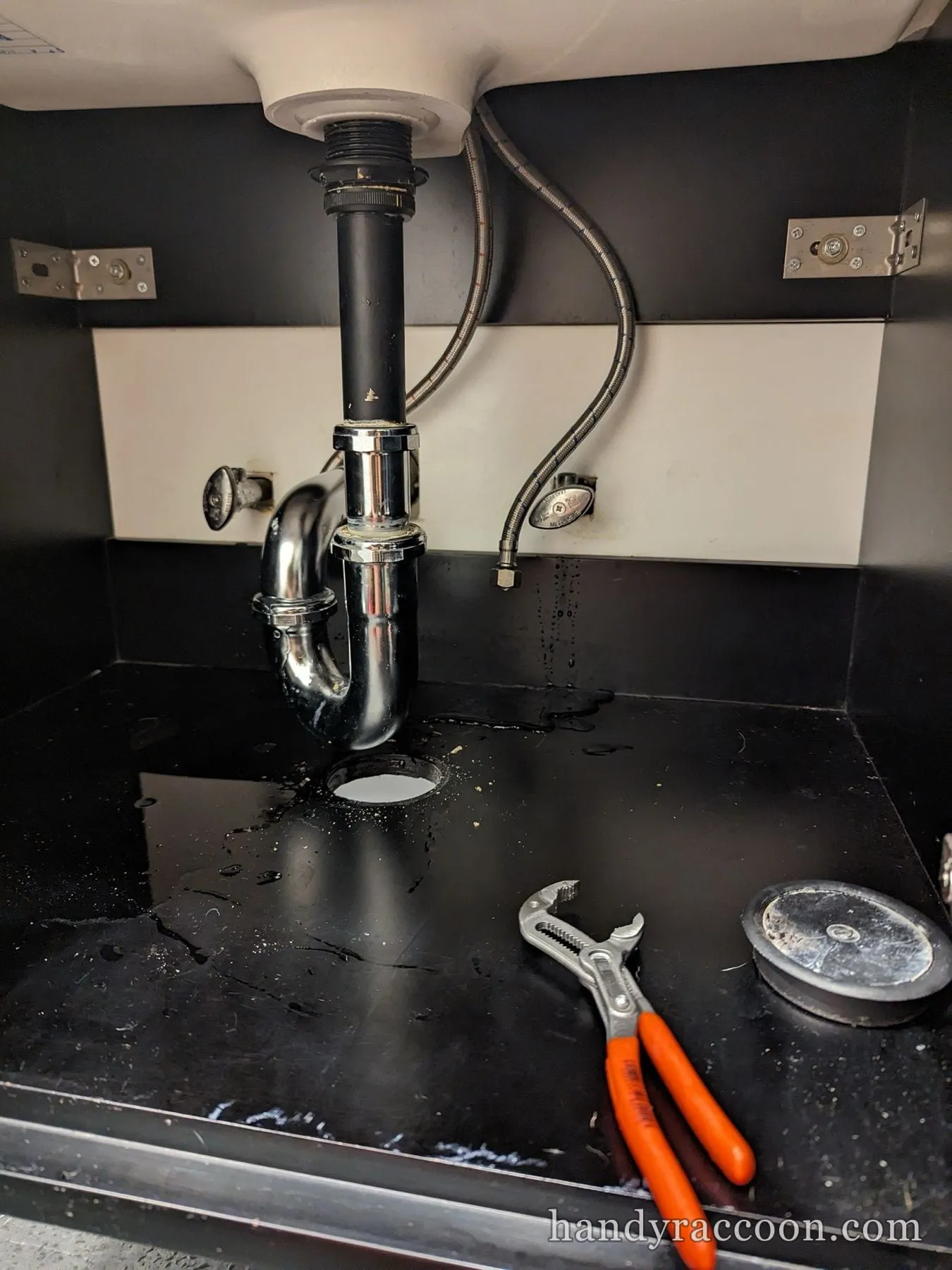 Chrome P-trap installed under a black vanity cabinet with pliers and some water on the base.