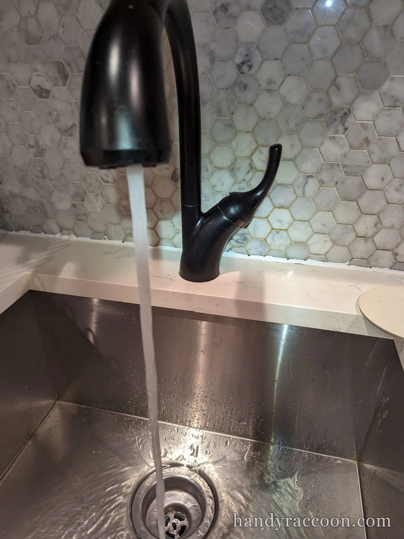 Black gooseneck kitchen faucet with water streaming into a stainless steel sink against a marble hex tile backsplash.